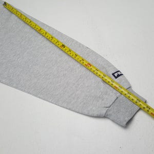 May include: A light grey sweatshirt sleeve with a dark grey ribbed cuff. The sleeve has a small navy blue patch with the brand name "FIL" in white. A yellow tape measure is laid across the sleeve, showing measurements in centimetres.
