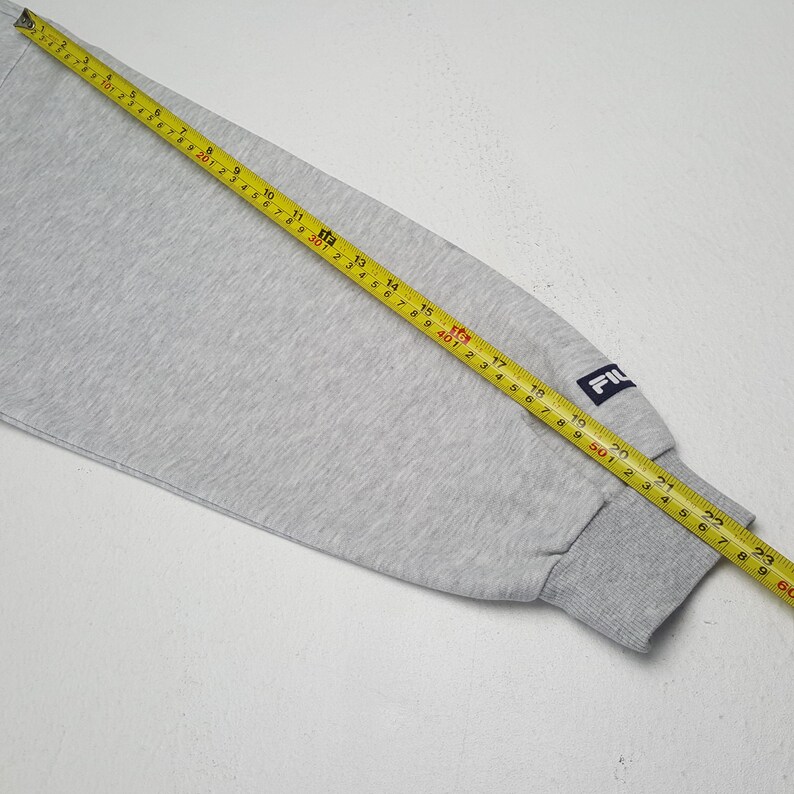 May include: A light grey sweatshirt sleeve with a dark grey ribbed cuff. The sleeve has a small navy blue patch with the brand name "FIL" in white. A yellow tape measure is laid across the sleeve, showing measurements in centimetres.