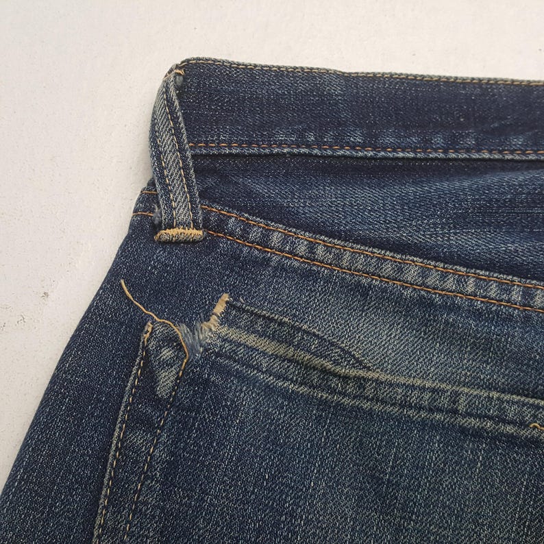 May include: A pair of dark blue denim jeans featuring a visible frayed edge on the corner of the back pocket. The fabric displays a classic twill weave texture with contrasting orange stitching along the seams and a single belt loop at the waist.