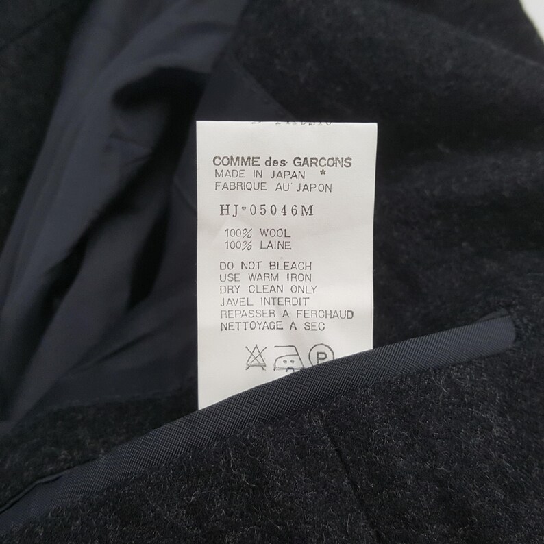 May include: Close-up of a dark gray wool jacket with a tag. The tag reads "COMME des GARCONS MADE IN JAPAN" and includes care instructions. The jacket is made of 100% wool.