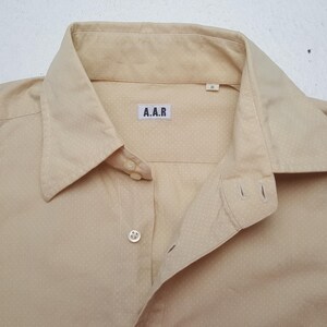 Pode incluir: Uma camisa de bot&otilde;es bege claro com um padr&atilde;o subtil de micro pontos brancos. A camisa tem um colarinho cl&aacute;ssico, um bot&atilde;o vis&iacute;vel na carcela frontal e uma etiqueta retangular branca no decote com o texto A.A.R e o tamanho M.