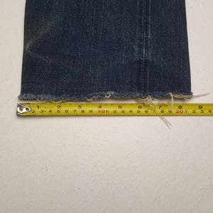 May include: A dark blue denim pant leg with a frayed, raw hem edge. A yellow measuring tape is positioned horizontally across the bottom of the fabric, showing a width measurement of approximately 9.5 inches from the side seam to the edge.