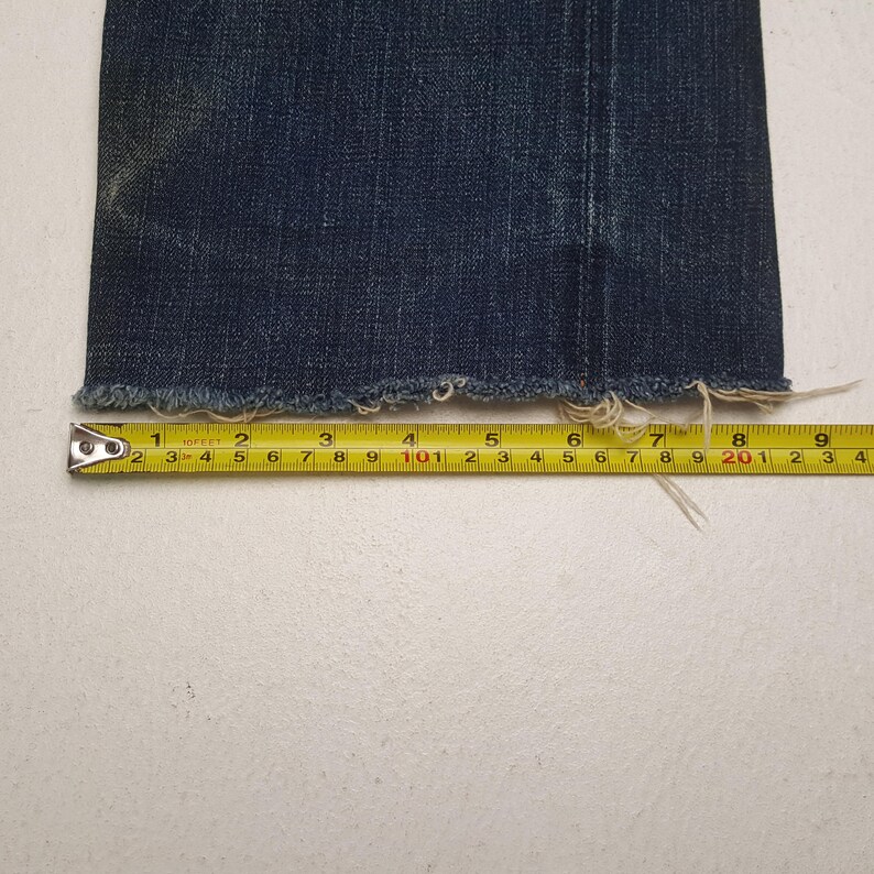 May include: A dark blue denim pant leg with a frayed, raw hem edge. A yellow measuring tape is positioned horizontally across the bottom of the fabric, showing a width measurement of approximately 9.5 inches from the side seam to the edge.