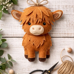 Puede incluir: Adorno hecho a mano de fieltro de una vaca de las Tierras Altas en tonos marrón y beige, con una cuerda de yute para colgar. La vaca tiene una melena desgreñada, orejas pequeñas y un diseño cosido. Tijeras negras y un ovillo de hilo.