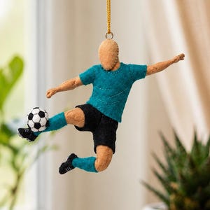 Puede incluir: Adorno de fieltro de un jugador de fútbol en pleno lanzamiento, suspendido de una cadena dorada. El jugador viste camiseta turquesa, pantalón corto negro y calcetines turquesa, con un balón de fútbol blanco y negro. El adorno cuelga de una rama.