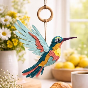 Peut inclure: Ornement en forme de colibri en feutre coloré, avec des accents bleus, turquoise, orange et jaunes, suspendu par un anneau en bois et une ficelle. L'oiseau a un long bec violet et est présenté dans un cadre lumineux et aéré. Un objet décoratif.