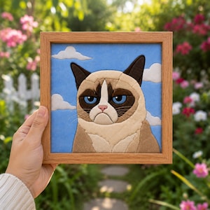 May include: A square fabric wall art piece in a light wood frame featuring a quilted applique of a grumpy cat with blue eyes and tan fur against a light blue sky background with white clouds. The cat has a distinct frown and dark brown ears.