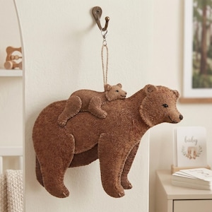 Op de afbeelding: Bruine vilten berendecoratie met een kleinere beer op zijn rug, hangend aan een haak. De beer heeft een getextureerd uiterlijk en is ontworpen voor de inrichting van een babykamer.