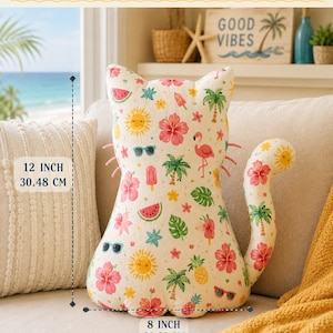 Könnte beinhalten: Ein katzenförmiges Plüschkissen aus cremefarbenem Stoff mit einem bunten Sommermuster aus Palmen, Flamingos, Hibiskusblüten, Ananas, Wassermelonen, Sonnenbrillen und Sonnen. Es ist 30,48 cm hoch und 20,32 cm breit.