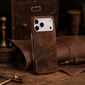Puede incluir: Una funda de cuero marrón oscuro para teléfono con una textura suave y mate. La funda presenta un recorte rectangular en la parte superior para acomodar un sistema de cámara de tres lentes y un flash, revelando la parte trasera blanca.