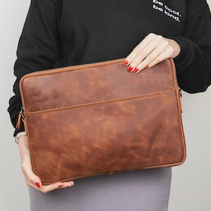 May include: A brown leather laptop sleeve with a zipper closure and a front pocket. The sleeve is rectangular and has a textured surface. The text "be cool. be kind." is visible on the upper left.
