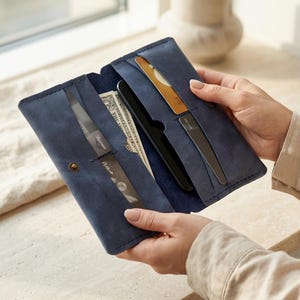 May include: A navy blue leather long wallet featuring multiple interior slots, currently holding several credit cards, a black smartphone, and paper currency. The wallet is constructed with visible stitching and a single metal snap closure.