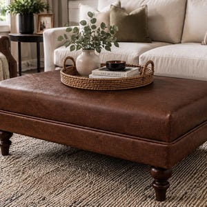 May include: A rectangular brown leather ottoman with a smooth surface and dark wooden turned legs. A round woven rattan tray sits on top, holding a white ceramic vase with green foliage, a small black candle, and a stack of two books.