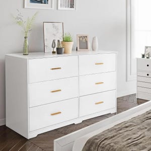 May include: A white six-drawer chest of drawers with gold-coloured handles. The furniture is in a bedroom, with decorative items on top, including plants, a vase, and picture frames. The room has a neutral colour palette.
