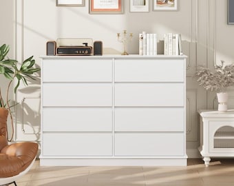 May include: A white dresser with eight drawers, a flat top, and a low base. A record player with speakers sits on top, along with books and decorative items. The dresser is in a room with framed artwork and a leather chair.