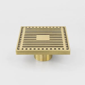 May include: A square-shaped brass floor drain featuring a series of horizontal parallel slats across the top surface and a solid central square section. The drain includes a cylindrical base component extending downward from the center of the grate.