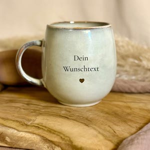 Könnte beinhalten: Eine hellgrüne Keramiktasse mit runder Form und Henkel. Die Tasse trägt den Text "Dein Wunschtext" und ein kleines Herzsymbol. Die Tasse steht auf einer Holzoberfläche.