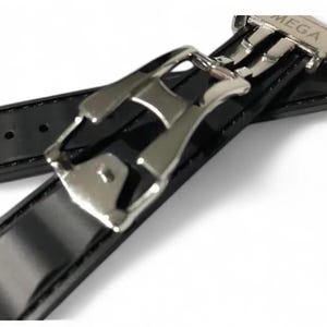 K&ouml;nnte beinhalten: Nahaufnahme eines schwarzen Uhrenarmbands mit gl&auml;nzendem Lackleder-Finish. Das Armband hat eine silberfarbene Schlie&szlig;e mit dem eingravierten Wort "MEGA". Das Armband ist auf Langlebigkeit und Stil ausgelegt.