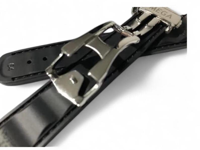 K&ouml;nnte beinhalten: Nahaufnahme eines schwarzen Uhrenarmbands mit gl&auml;nzendem Lackleder-Finish. Das Armband hat eine silberfarbene Schlie&szlig;e mit dem eingravierten Wort "MEGA". Das Armband ist auf Langlebigkeit und Stil ausgelegt.