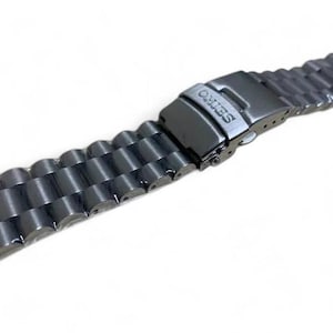 May include: A dark gray metal watch band with a deployment clasp. The band is made of connected, rounded links. The clasp is marked with the brand name "SEIKO".