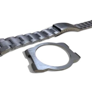 Könnte beinhalten: Ein silberfarbenes Uhrenarmband und eine Uhrengehäusekomponente werden vor weißem Hintergrund gezeigt. Das Armband besteht aus verbundenen Metallgliedern. Die Uhrengehäusekomponente ist kreisförmig mit einem Ausschnitt für das Zifferblatt.