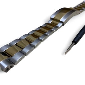 Puede incluir: Una correa de reloj bicolor con eslabones plateados y dorados, y una herramienta con mango negro y punta plateada. La correa del reloj es de metal y tiene un acabado pulido. La herramienta es probablemente para ajustar o reparar la correa.