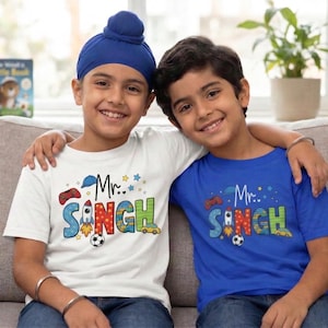 May include: Two young boys wearing matching t-shirts with the text "Mr. Singh" in colorful letters. One shirt is white, the other blue. Both shirts feature a rocket, soccer ball, and other graphics. The boys are smiling.