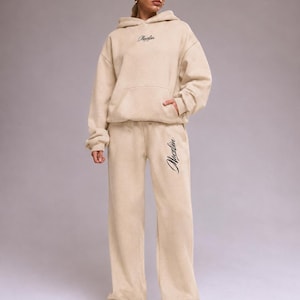 Puede incluir: Un conjunto beige a juego compuesto por una sudadera con capucha de manga larga y pantalones de chándal anchos. La sudadera tiene un bolsillo canguro y el texto negro "Macelin" en el pecho. El pantalón lleva el texto negro "Macelin" en la pierna.