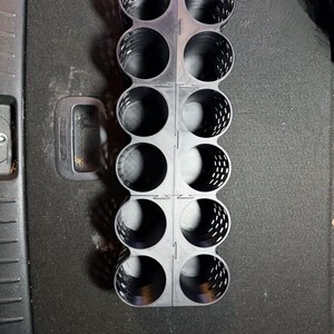 May include: A black plastic organizer with multiple circular compartments. The organizer is designed to hold cylindrical objects, with a grid-like structure and a handle. The design features a series of perforated holes for ventilation.