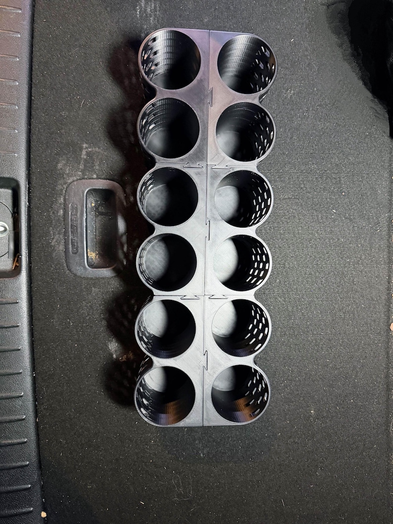 May include: A black plastic organizer with multiple circular compartments. The organizer is designed to hold cylindrical objects, with a grid-like structure and a handle. The design features a series of perforated holes for ventilation.