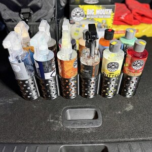 May include: A collection of car detailing products, including spray bottles and bottles of liquid, are organized in a metal holder. The products are various colors and brands, with labels visible. The holder is in a car trunk.
