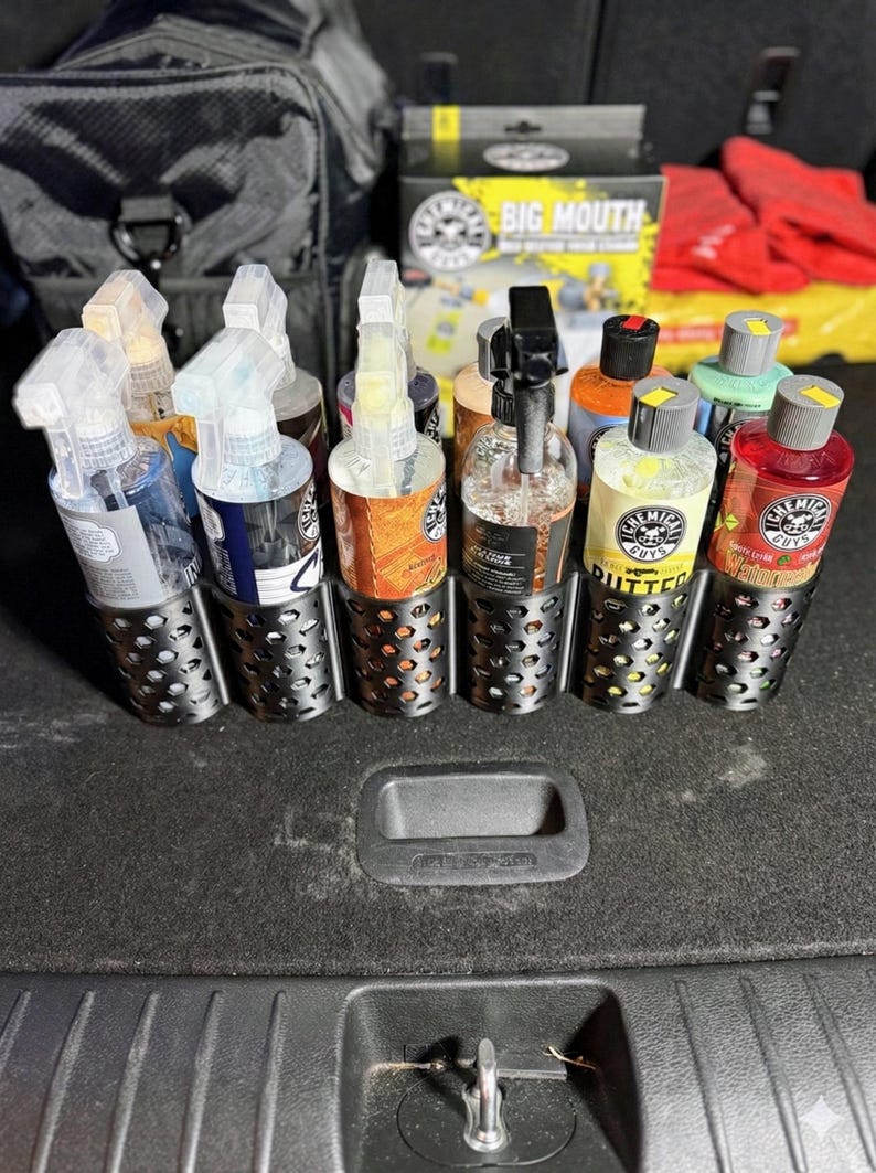 May include: A collection of car detailing products, including spray bottles and bottles of liquid, are organized in a metal holder. The products are various colors and brands, with labels visible. The holder is in a car trunk.