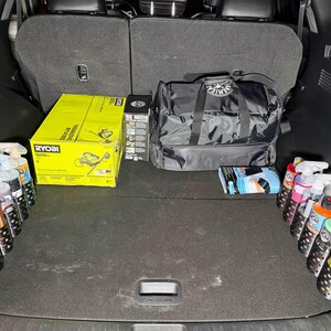 May include: A car trunk filled with detailing supplies. Includes a yellow Ryobi box, a black duffel bag, spray bottles, and other detailing products. The trunk is organized with products in holders.