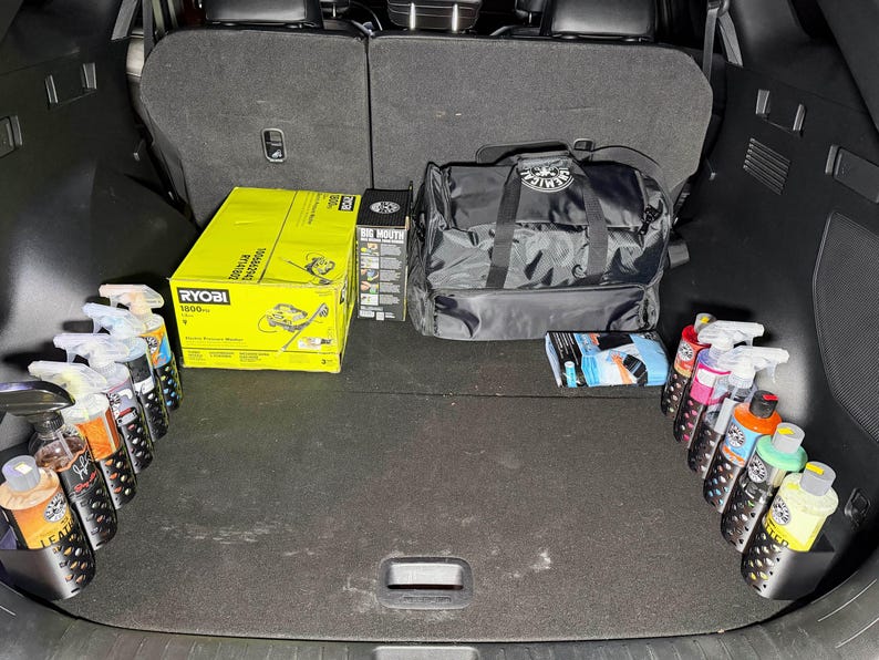 May include: A car trunk filled with detailing supplies. Includes a yellow Ryobi box, a black duffel bag, spray bottles, and other detailing products. The trunk is organized with products in holders.