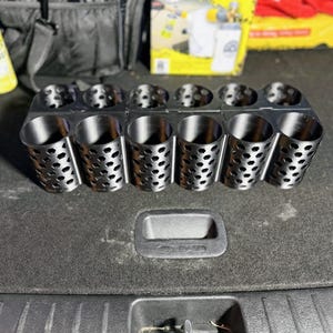 May include: A black metal organizer with multiple cylindrical compartments. Each compartment has a perforated design. The organizer is designed to hold various items, likely for automotive detailing or storage. The Big Mouth product box is visible in the background.
