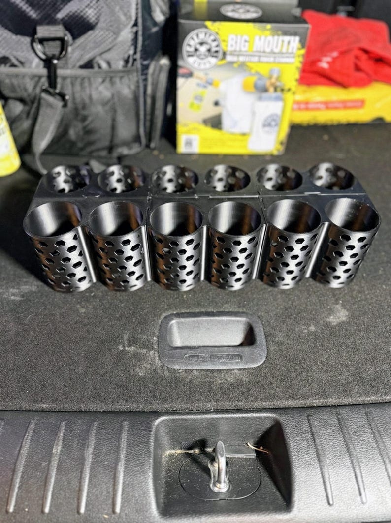 May include: A black metal organizer with multiple cylindrical compartments. Each compartment has a perforated design. The organizer is designed to hold various items, likely for automotive detailing or storage. The Big Mouth product box is visible in the background.