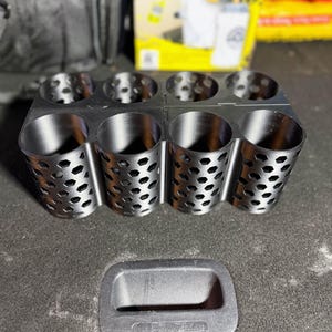 May include: A black metal organizer with eight cylindrical compartments, each with a honeycomb pattern. A black rectangular handle is below the organizer. The background includes a box with the words "BIG MOUTH" and a car detailing product.