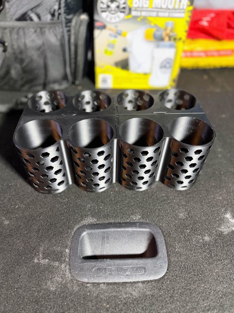 May include: A black metal organizer with eight cylindrical compartments, each with a honeycomb pattern. A black rectangular handle is below the organizer. The background includes a box with the words "BIG MOUTH" and a car detailing product.