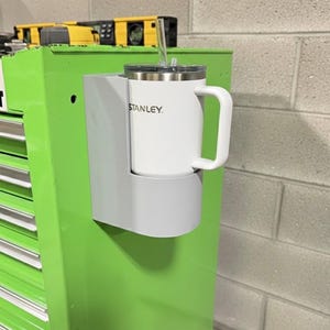 May include: A white Stanley mug with a handle and a silver lid, held in a gray cup holder attached to a green toolbox. The mug has the Stanley logo printed on it. A clear straw is in the mug.