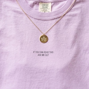 May include: A light purple t-shirt with the text "IF YOU CAN READ THIS ASK ME OUT" printed in black. A gold-colored necklace with a round pendant that reads "NEW BALTIMORE, MICHIGAN EST. 2026" is on the shirt.