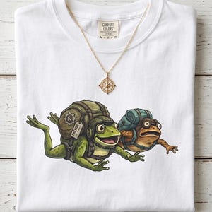 May include: White Comfort Colors t-shirt featuring a cartoon illustration of two frogs with backpacks. One frog is green, the other is brown. A gold necklace with a compass pendant is also visible.