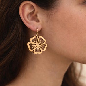 May include: Gold-colored flower-shaped earrings. The earrings are worn on a person's ear. The earrings have a cutout design, with a hook closure. The earrings are a warm gold color.