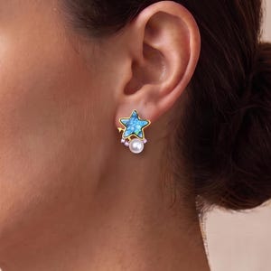 May include: A gold-toned stud earring shaped like a five-pointed star with a blue marbled inlay. The earring features a small white pearl dangling from the bottom, accented by three tiny clear stones set along the gold frame near the pearl.