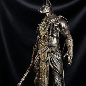 Peut inclure: Statue de bronze d'Anubis, dieu égyptien de l'au-delà, avec une tête de chacal et des détails ornés. La statue tient une lance et se dresse sur un piédestal rond. La figure est musclée et porte une armure détaillée.