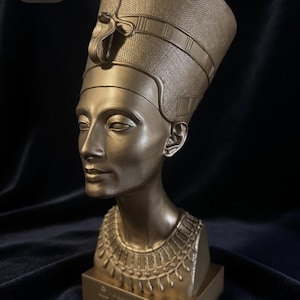 Pode incluir: Busto dourado de Nefertiti, com aproximadamente 18 cm de altura, com detalhes intrincados na touca e no colar. O busto é exibido em uma base retangular. A imagem inclui o texto "18 cm".