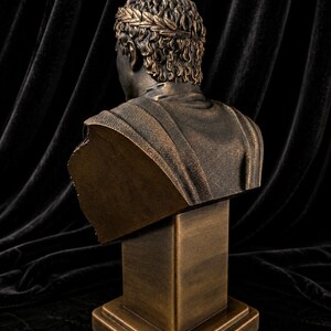 Pode incluir: Busto em bronze de um imperador romano, visto por tr&aacute;s. A escultura apresenta uma coroa de louros e texturas detalhadas no cabelo e nas roupas. O busto &eacute; montado em um pedestal quadrado.