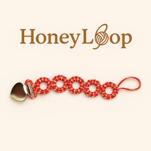 Puede incluir: Un clip para chupete con un cierre metálico en forma de corazón y cinco anillos a rayas rojas y blancas. El clip está sujeto a un cordón rojo. El logotipo "HoneyLoop" está en la parte superior.