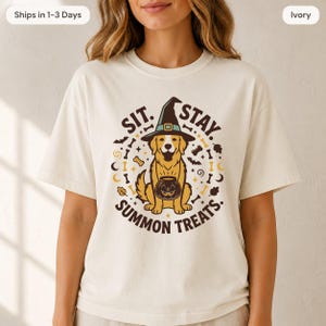 May include: An ivory short-sleeve t-shirt featuring a graphic of a golden retriever wearing a black witch hat and holding a jack-o'-lantern bucket. The text reads SIT. STAY. SUMMON TREATS. surrounded by small icons of bones, bats, and leaves.