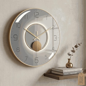 May include: A modern wall clock with a gray face, gold-tone hands, and a gold-colored pendulum. The clock has a gold-colored frame and black numerals. The clock is a decorative item for a home or office.