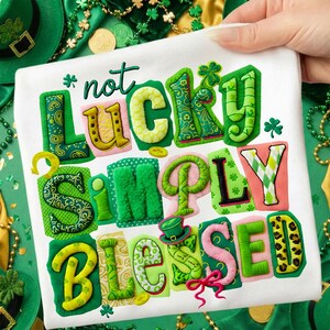 May include: White t-shirt with the embroidered phrase "not LUCKY simply BLESSED" in various colors and patterns. The design includes shamrocks, a leprechaun hat, and gold coins, all set against a green and gold background, perfect for St. Patrick's Day.